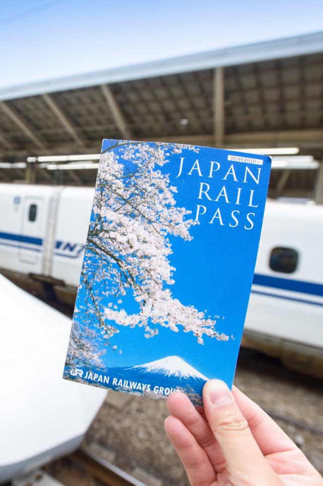 Hånden holder en Japan Rail Pass-billett foran en Shinkansen på Tokyo stasjon