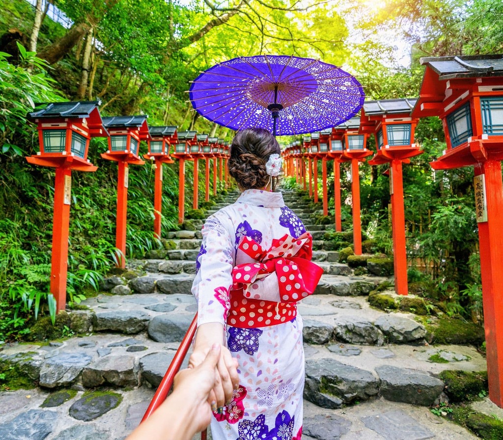Kvinne i kimono går opp steintrapp med røde lykter i Japan.