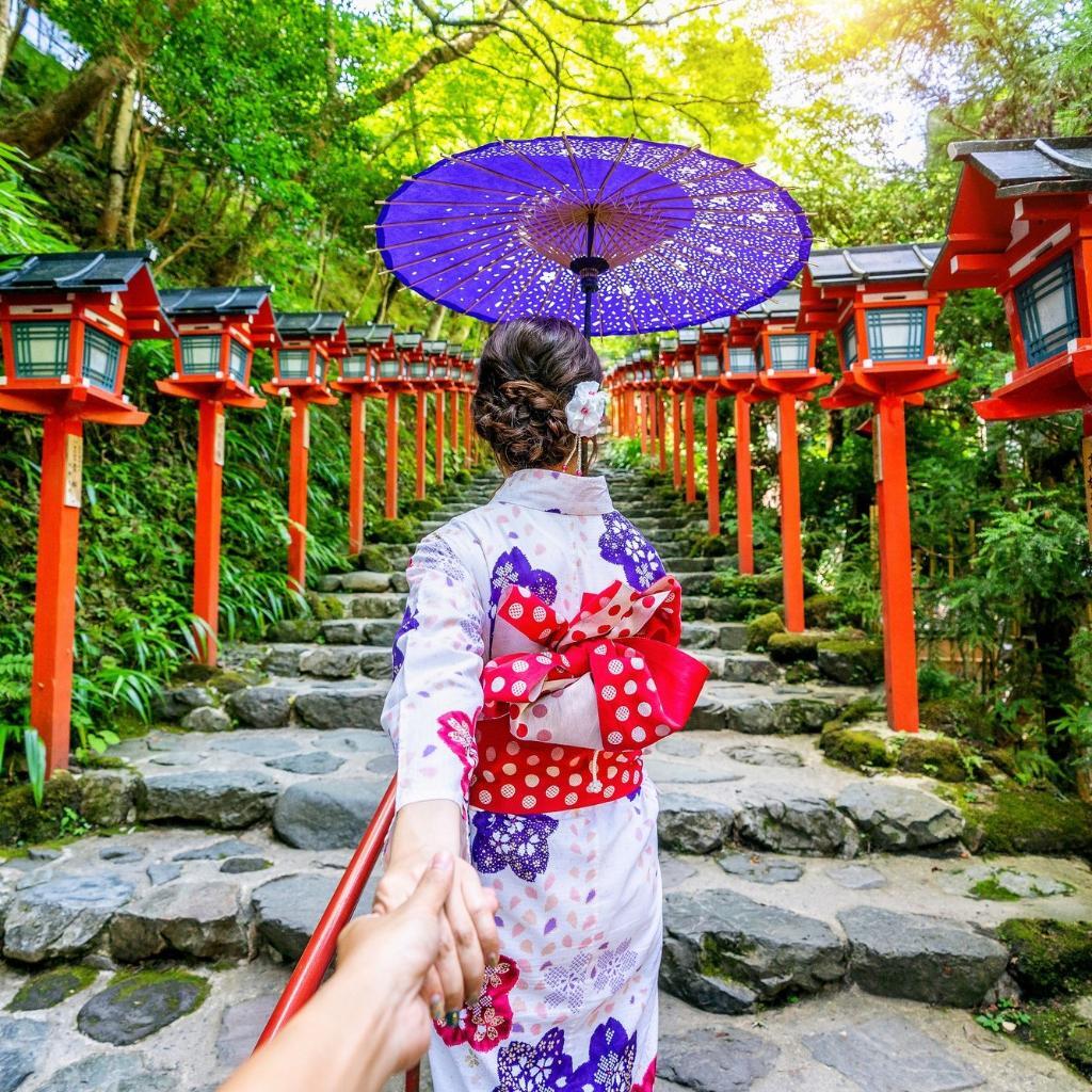 Kvinne i kimono går opp steintrapp med røde lykter i Japan.
